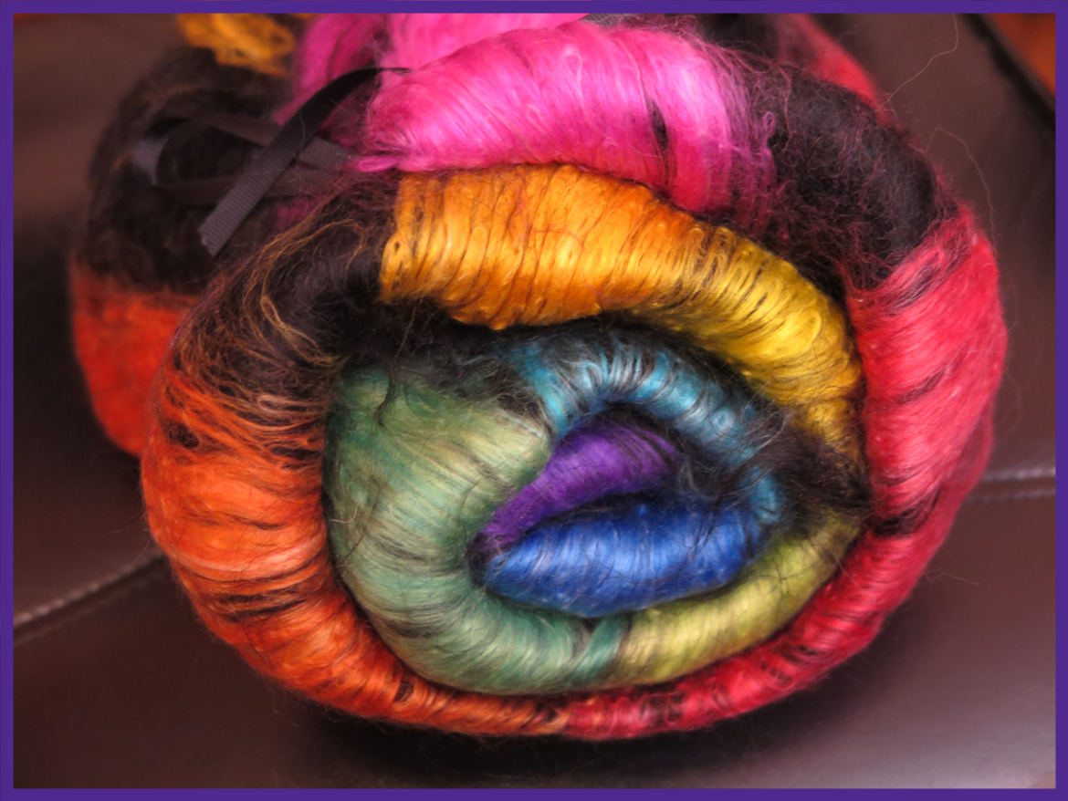A batt of black alpaca with a layer of silk in a rainbow from purple through to pick is rolled up and tied together with a black ribbon.