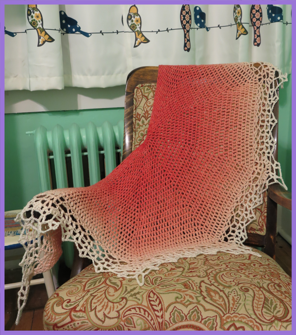 A peach-pink to white gradient crochet lace shawl is draped over a wood rocking chair with a rust red and green paisley pattern. The shawl contrasts with the mint-green paint of the wall behind.