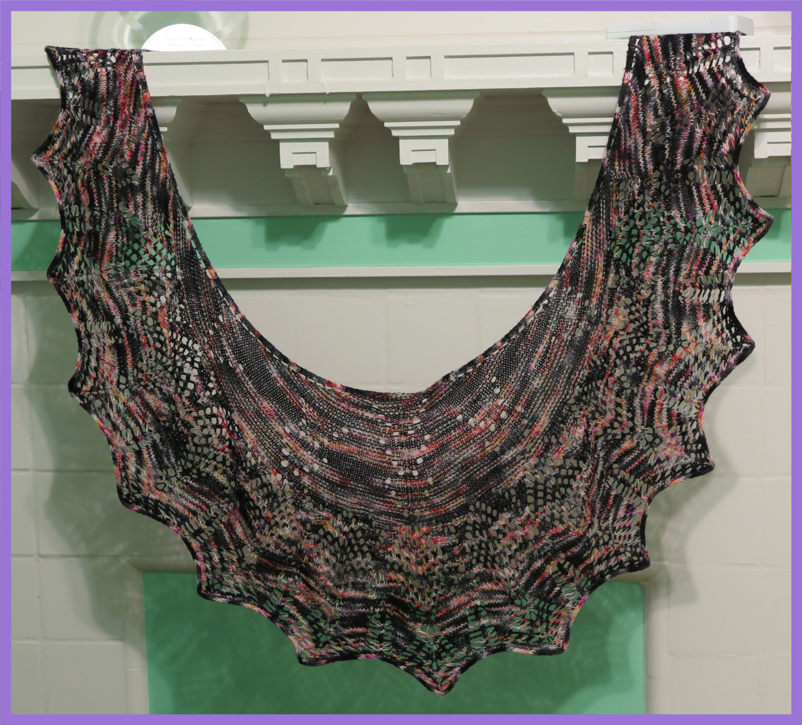 A lace shawl is hung from a mantle of a filled in fireplace painted in white and mint green. The yarn is black with splashes of pinks, oranges, white and grey and the lace pattern forms arrow heads and fletching.