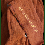 An orange sweater sleeve, not quite finished, with the words "all of this must go" written across it.