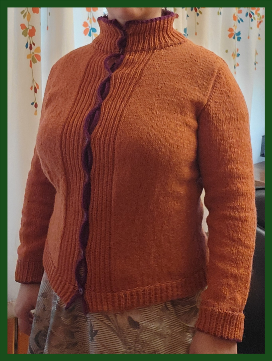 Jayebird wears her Nithya sweater knit in orange with contrast details in a reddish purple. It buttons down the front with ribbing along the front and a tall collar.